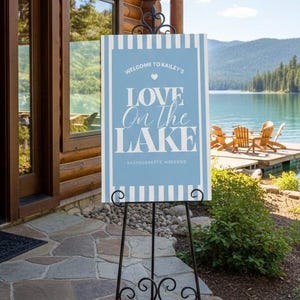 Könnte beinhalten: Ein Willkommensschild auf einer schwarzen Metalleasel. Das Schild ist hellblau mit weißem Text: "Welcome to Kailey's Love on the Lake Bachelorette Weekend." Der Hintergrund zeigt einen See und einen Holzsteg.