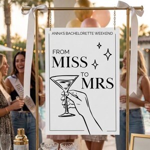Puede incluir: Un letrero de metal dorado con un letrero blanco que dice "Anna's Bachelorette Weekend" y "From Miss to Mrs" con una ilustración de copa de martini. Cintas blancas y cadenas doradas están unidas al letrero. El letrero está sobre una mesa de madera con cristalería.