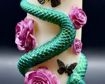 May include: Ivory pillar candle decorated with a teal and green snake, pink roses, and black butterflies. The snake wraps around the candle, and the roses and butterflies are scattered across the surface.