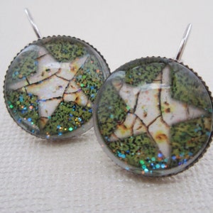 May include: A pair of round earrings with a star design. The earrings feature a white star with a cracked pattern set against a green background with glitter accents. The earrings have a silver-colored hook.