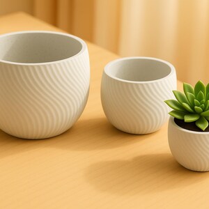 Peut inclure: Trois pots de fleurs en céramique blanche avec un motif ondulé. Un pot contient une plante succulente verte. Les pots sont de tailles différentes et reposent sur une surface brun clair. Le fond est jaune clair.