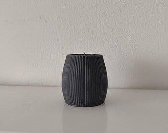 May include: A black, cylindrical candle with vertical ribbed detailing. The candle sits on a white surface against a white wall. The candle has a wick at the top.