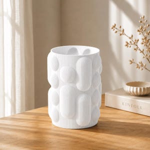 Peut inclure: Un vase cylindrique blanc présentant une surface texturée avec des protubérances arrondies en forme de pilules. L'extérieur du vase est entièrement recouvert de fines nervures verticales qui suivent les contours des formes géométriques.
