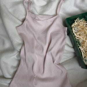May include: A light pink ribbed tank top with adjustable straps. The top is made of a soft, stretchy material and has a scoop neckline. The tank top is laying on a white, textured surface.