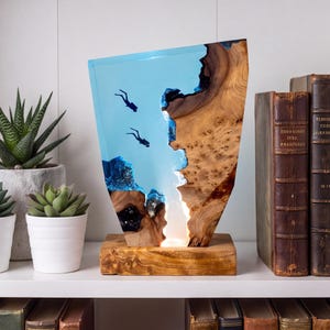 Könnte beinhalten: Eine dekorative Lampe aus einer natürlichen Holzscheibe, die in durchscheinendes blaues Harz eingearbeitet ist. Der Harzbereich enthält zwei kleine Silhouetten von Tauchern. Das Stück steht auf einem rechteckigen Holzsockel und strahlt warmes Licht aus.