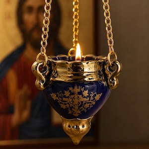 Peut inclure: Une lampe à huile suspendue composée d'un bol en céramique bleu profond orné de motifs floraux et de croix dorés. La lampe présente un bord et une base en métal doré, suspendue par trois chaînes métalliques dorées. Une flamme brûle à l'intérieur.