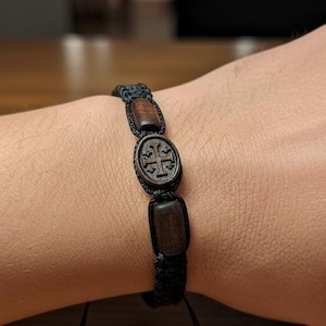 Puede incluir: Pulsera de macramé negro con cuentas de madera. La pulsera presenta una cuenta central ovalada con un diseño de cruz tallada, flanqueada por dos cuentas rectangulares de madera. La pulsera es ajustable con nudos corredizos.