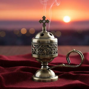 Peut inclure: Un brûleur d'encens en bronze avec une croix sur le dessus, dégageant de la fumée. Le brûleur est posé sur un tissu rouge foncé, avec un coucher de soleil en arrière-plan. Le brûleur présente des détails floraux.