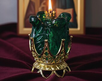 May include: A lit green glass candle with a gold crown-shaped holder and a gold base. The candle is in front of a religious icon with a blurred image of a man. The candle is on a burgundy surface.