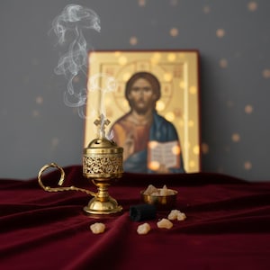 May include: A gold-colored censer with intricate floral designs, emitting wisps of smoke, sits on a deep red velvet surface. A small bowl of incense and an icon of Jesus are in the background. The censer has a cross on top.