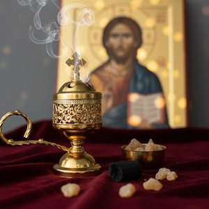 May include: A gold-colored censer with a cross on top, emitting wisps of smoke. The censer has a floral design and a handle. Beside it are a small bowl with amber-colored resin, a piece of charcoal, and an icon of Jesus.