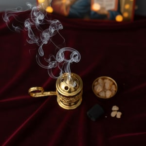 May include: A gold-colored incense burner with a handle, emitting wisps of white smoke. Beside it are a small bowl of incense, a black charcoal disc, and loose incense pieces. The items rest on a deep red velvet surface.