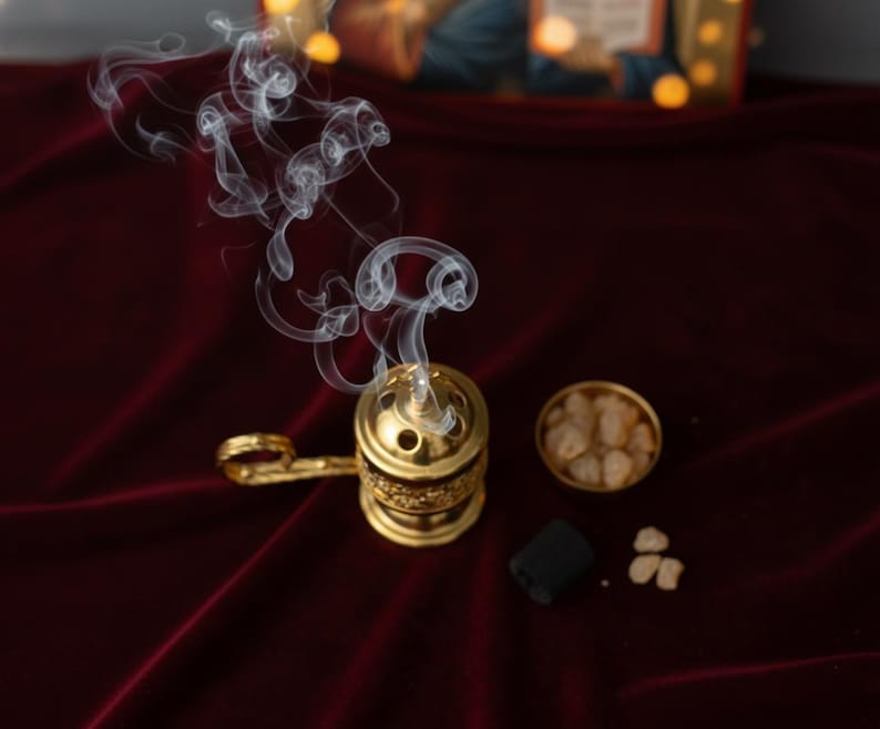 May include: A gold-colored incense burner with a handle, emitting wisps of white smoke. Beside it are a small bowl of incense, a black charcoal disc, and loose incense pieces. The items rest on a deep red velvet surface.