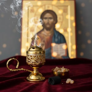 May include: A gold-colored censer with floral detailing, emitting wisps of smoke, sits on a burgundy velvet surface. A small gold bowl holds amber-colored resin, and a black charcoal piece is nearby. An icon of Jesus is in the background.