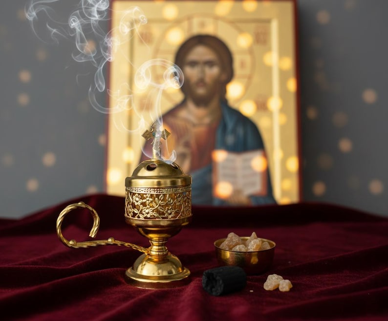 May include: A gold-colored censer with floral detailing, emitting wisps of smoke, sits on a burgundy velvet surface. A small gold bowl holds amber-colored resin, and a black charcoal piece is nearby. An icon of Jesus is in the background.