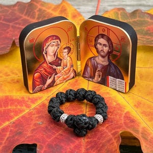 May include: A black prayer rope with silver accents rests on a colorful leaf, next to a small, hinged icon depicting religious figures. The icon has gold trim and detailed artwork.