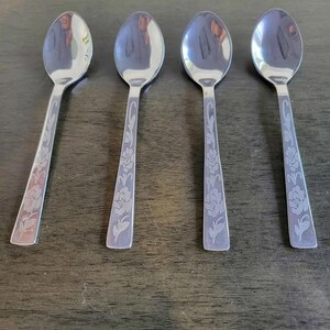 May include: Four stainless steel spoons with floral etched handles. The spoons have a reflective surface and are arranged on a dark wooden surface. The spoons are designed for serving or eating.