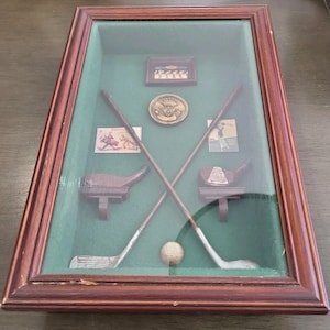 May include: A rectangular wooden shadow box frame with a green felt backing containing two crossed miniature golf clubs, a golf ball, a circular metallic medallion, and three small rectangular cards featuring vintage golf illustrations.