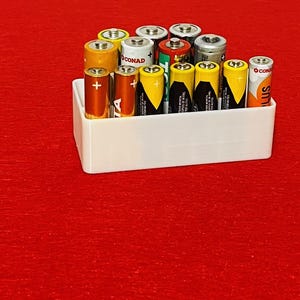May include: A white rectangular plastic storage container filled with an assortment of upright cylindrical batteries. The batteries feature various colors including orange, yellow, silver, and red, with visible plus and minus polarity symbols.