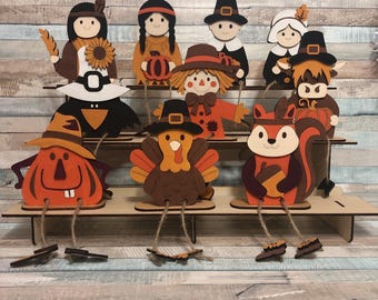 May include: A selection of wooden Thanksgiving-themed figurines. The figurines include a Native American, a pilgrim, a scarecrow, a squirrel, a turkey, and a pumpkin. Each figure is painted in autumnal colours and has dangling legs.