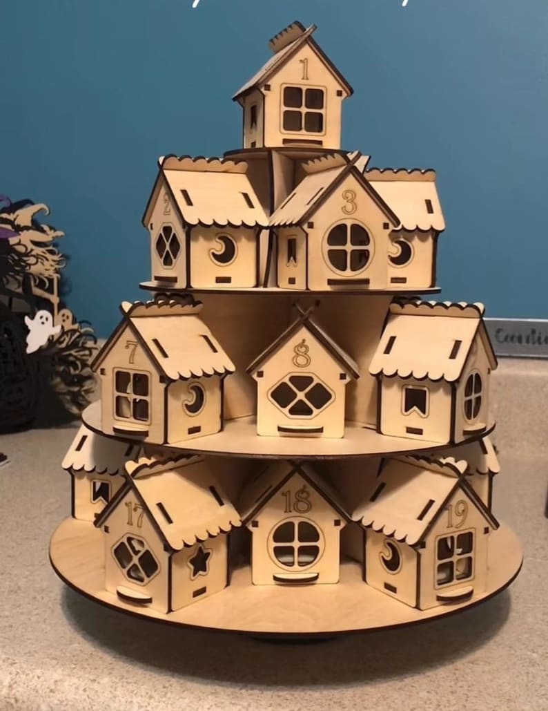 May include: A multi-tiered wooden advent calendar in the shape of a village. The natural wood houses are numbered and feature different window designs. The houses are arranged on circular platforms, forming a multi-level display.