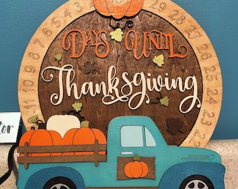 May include: Wooden Thanksgiving countdown sign. A blue truck carries orange and white pumpkins. The sign has a circular background with the words "Days Until Thanksgiving" and a pumpkin at the top. Numbers 1-32 are around the circle.
