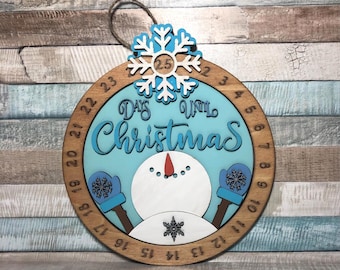 May include: Wooden Christmas countdown ornament. The round ornament features a snowman design with the words "Days Until Christmas" in blue. A snowflake with the number 25 is at the top, and the days are numbered around the edge.