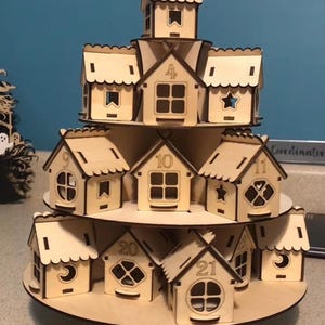 May include: A tiered wooden advent calendar in the shape of a village. The natural wood houses are numbered and have various window cutouts, including stars and moons. The calendar is designed to hold small treats or gifts.