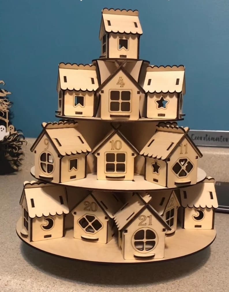 May include: A tiered wooden advent calendar in the shape of a village. The natural wood houses are numbered and have various window cutouts, including stars and moons. The calendar is designed to hold small treats or gifts.