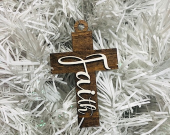 Könnte beinhalten: Ein hölzernes Kreuzornament in Dunkelbraun mit dem Wort "Faith" in Schreibschrift. Das Kreuz hat ein Loch zum Aufhängen. Der Schmuck wird vor einem weißen, strukturierten Hintergrund, möglicherweise einem Weihnachtsbaum, präsentiert.