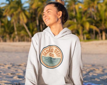 May include: A light gray hoodie with a front pocket and a circular logo that reads "Faded Reef Co." in a retro design. The hoodie is worn by a person sitting on a beach with palm trees in the background.