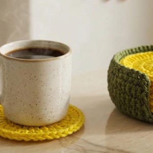 K&ouml;nnte beinhalten: Eine beige Keramiktasse mit dunklem Kaffee steht auf einem gelben geh&auml;kelten Untersetzer. Ein Satz gelber Untersetzer befindet sich in einem gr&uuml;nen geh&auml;kelten Halter. Die Untersetzer und der Halter sind handgefertigt.