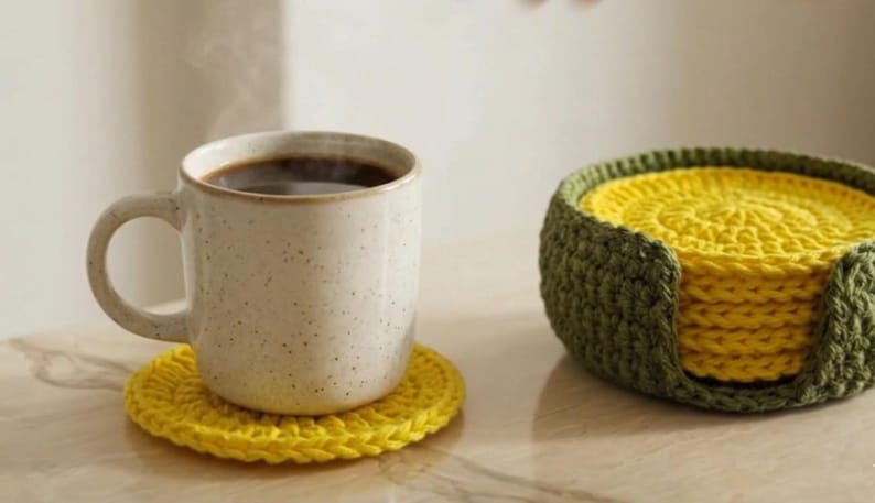 K&ouml;nnte beinhalten: Eine beige Keramiktasse mit dunklem Kaffee steht auf einem gelben geh&auml;kelten Untersetzer. Ein Satz gelber Untersetzer befindet sich in einem gr&uuml;nen geh&auml;kelten Halter. Die Untersetzer und der Halter sind handgefertigt.
