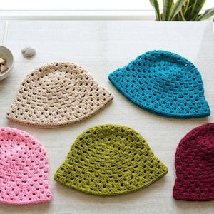May include: Five crochet bucket hats in various colors are displayed on a white table. The hats are in beige, turquoise, pink, olive green, and burgundy. The hats have a breathable, open weave design, perfect for warm weather.