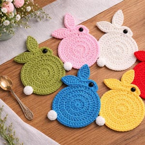 Op de afbeelding: Een verzameling van zes kleurrijke, gehaakte konijnvormige onderzetters. De onderzetters zijn in groen, roze, wit, blauw, geel en rood. Elke onderzetter heeft een zwart knoopoog en een witte pomponstaart.