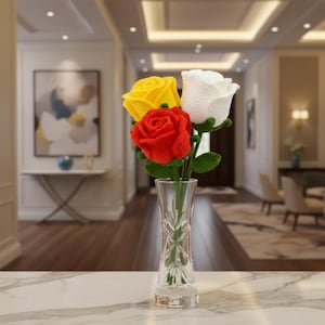 May include: A clear glass vase holding a bouquet of three crocheted roses. The roses are in vibrant colors: red, yellow, and white. The vase is placed on a marble surface, with a blurred background of a well-lit room.
