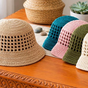 May include: A collection of five crochet bucket hats in beige, teal, light pink, olive green, and cream. Each hat features a solid crown and brim with a square-patterned openwork crochet design around the midsection, showcasing a textured finish.