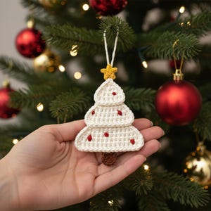May include: A handmade crochet Christmas tree ornament. The tree is white with red accents and a yellow star on top. The tree is suspended by a white string and has a brown trunk. The ornament is displayed in front of a Christmas tree with red and gold ornaments.