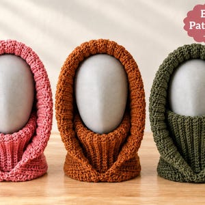 May include: Three knitted balaclava hoods in pink, rust orange, and olive green, each displayed on a gray mannequin head. The items feature a ribbed texture and a wide opening around the face. A pink label in the corner reads English Pattern PDF.