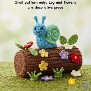 May include: A blue and green crocheted snail toy with black eyes and a smiling mouth. The text above states: Snail pattern only. Log and flowers are decorative props. The snail is positioned on a brown crocheted log with small flowers.