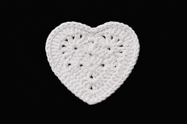 May include: A white, crocheted heart-shaped item against a black background. The heart is made of white yarn, with a textured surface and visible stitch patterns. The edges are neatly defined, and the overall shape is symmetrical.