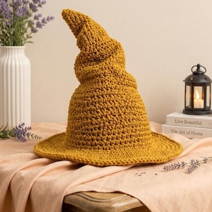 May include: A mustard yellow crocheted hat with a wide brim and a tall, pointed, slightly slumped crown. The hat is constructed from thick yarn with a visible textured stitch pattern, creating a structured yet flexible form for the head covering.