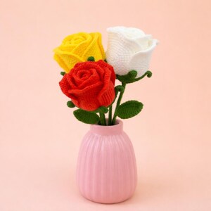 May include: A pink vase holds three crocheted roses: one yellow, one red, and one white. The roses have green stems and leaves. The vase has a ribbed design and sits against a soft pink background.