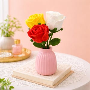May include: A small pink vase holding three crocheted roses: one yellow, one red, and one white. The vase sits on a closed book. Green leaves and stems support the flowers. The background is a soft pink.