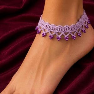 May include: A lavender crochet anklet featuring a scalloped edge design with small, matching purple beads dangling from each point of the lace pattern. The intricate thread work creates a delicate, decorative band that encircles the ankle area.