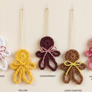May include: Five crocheted gingerbread-style ornaments in white, yellow, dark brown, light brown, and pink. Each figure features a small bow and a hanging loop. Text labels include THE CLASSIC, GINGERBREAD, YELLOW, LEMD-CRAFTED, and HAND-COLER TWINE.