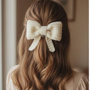 May include: A cream-colored, crocheted bow hair clip is secured in the hair. The bow is centered at the back of the head, with long, wavy brown hair cascading down. The clip has a silver-colored bar.