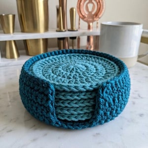 May include: A set of six handmade crochet coasters in shades of teal and blue, held in a matching holder. The coasters are circular with a spiral design, perfect for protecting surfaces from drink condensation. The holder has a sturdy, rounded shape.