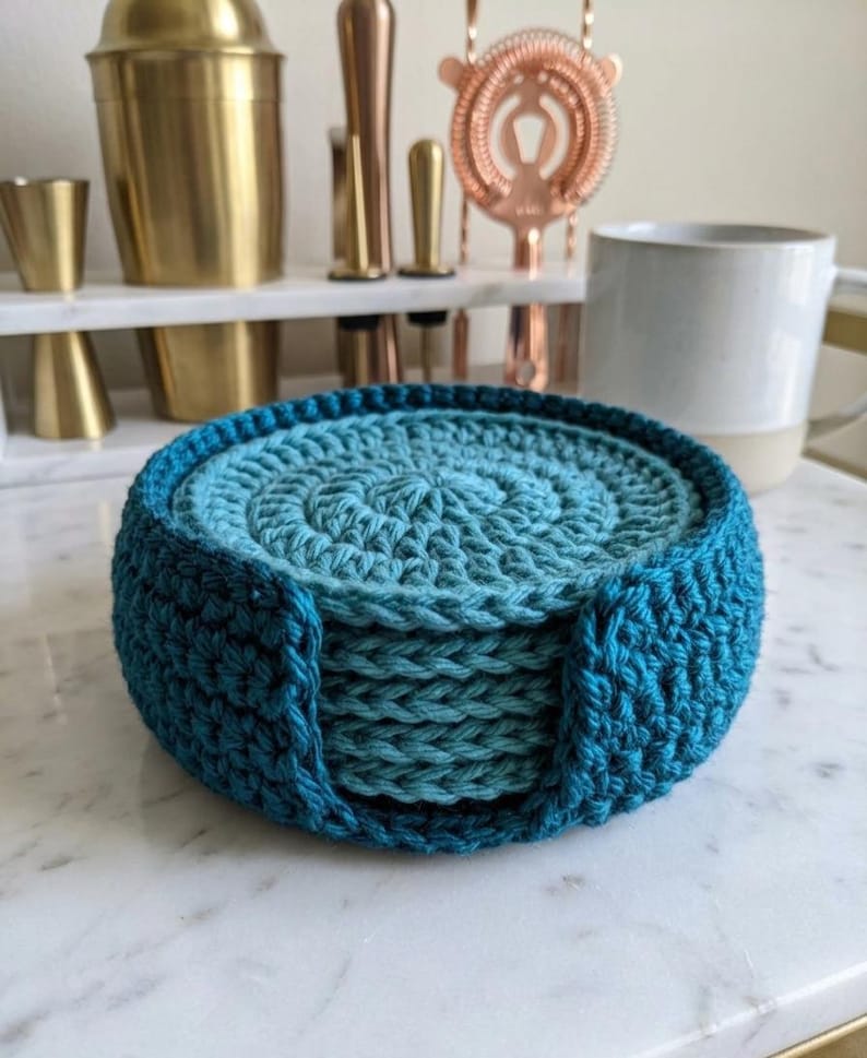 K&ouml;nnte beinhalten: Ein Set aus sechs handgeh&auml;kelten Untersetzern in T&uuml;rkis- und Blaut&ouml;nen, gehalten in einem passenden Halter. Die Untersetzer sind kreisf&ouml;rmig mit einem Spiraldesign, ideal zum Schutz von Oberfl&auml;chen vor Kondenswasser.