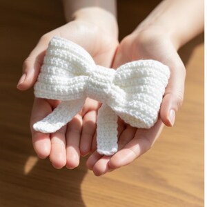 May include: A handmade, white crochet bow. The bow is held in open hands, showcasing its textured, knotted design. The bow has two loops and two long tails, perfect for hair accessories or decorative purposes.